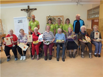 Acht Senioren sitzen nebeneinander auf Stühlen und präsentieren ihre Urkunden. Dahinter stehen Betreuungskräfte und der Caritasvorstand