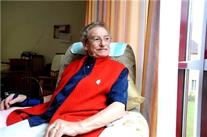 Eine ältere Frau in blauer Bluse und roter Jacke sitzt in ihrem Zimmer in einer Altenpflegeeinrichtung und schaut lächelnd aus dem Fenster. 