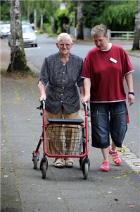 Eine Mitarbeiterin begleitet eine Bewohnerin eines Caritas-Altenzentrums bei einem Spaziergang. Die Dame ist auf einen Rollator angewiesen. 