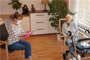 Eine Dame mit Mundschutz sitzt auf einem Stuhl und blättert in einem Heft. Ihr gegenüber sitzt eine ältere Dame. Vor Ihr steht Ihr Rollator. 