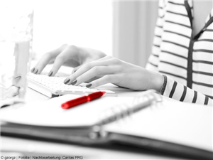 Bildtotale: Frauenhände schreiben auf der Computertastatur. In Graustufen. Im Vordergrund rot hervorgehobener Stift. (c) gzorgz|fotolia.de|Nachberarbeitung: Caritas FRG.