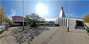 Öffentlicher Platz in grellem Sonnenschein, im Hintergrund ein moderner Kirchbau mit Nebengebäuden