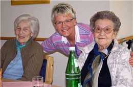 Porträtfoto von zwei Seniorinnen, umarmt von einer herzlich lachenden Pflegefachkraft 