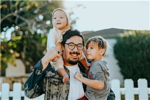 Ein dunkelhaariger Mann mit Brille und Bart lächelt in die Kamera und hält ein Kleinkind auf dem Arm und ein Baby auf den Schultern