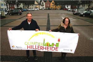 099-2017_Quartiersmanagement_Hillerheide die Quartiersmanager Monika Wagner-van der Straten und Björn Schmidt-Freistühler stehen auf dem Gertrudisplatz in Hillerheide und halten ein Banner mit der aufschrift 'Quartier Hillerheide'.