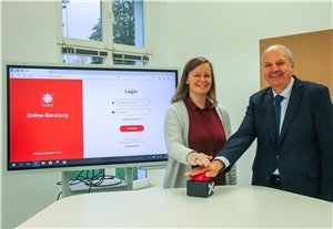 Das Foto zeigt zwei Menschen, die auf einen roten Knopf drücken, im Hintergrund wird auf einem großen Bildschirm die Eingangsseite der Online-Beratung gezeigt.