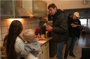Das Foto zeigt zwei Männer und eine junge Frau mit Baby auf dem Arm in einer Küche.