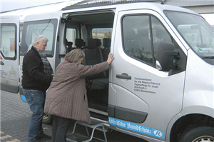 Ein älterer Mann hilft einer älteren Frau beim Einstieg in einen Kleintransporter, vor dem eine Trittleiter als Einstiegshilfe steht