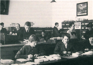 Historische Aufnahme in Schwarz-Weiß: Einige weibliche und männliche Mitarbeiter stehen bzw. sitzen in einem Büroraum mit Schreibbänken und Regalen und arbeiten
