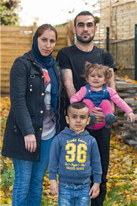 Eine Flüchtlingsfamilie bestehend aus einem Elternpaar, einem Jungen und einem Mädchen, stehen zusammen in einem Garten. Der Mann trägt das Mädchen in seinen Armen.
