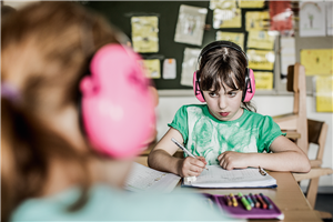 Eine Schülerin mit einem pinken Gehörschutz sitzt an einem Tisch in einem Klassenzimmer und erledigt Schulaufgaben. Am linken Bildrand ist ein weiteres Kind mit pinkem Gehörschutz zu sehen.