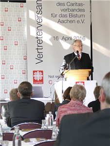 Prof. em. Klaus J. Bade steht an einem Pult und hält eine Rede. Im Vordergrund sind einige Teilnehmer der Vertreterversammlung des DiCV Aachen zu sehen.