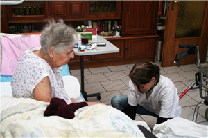 Eine Seniorin sitzt in einem Wohnraumraum auf ihrem Krankenbett. Vor ihr kniet eine junge Pflegerin und hantiert an ihren Füßen.