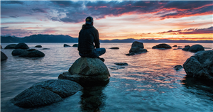Ein Jugendlicher sitzt im Schneidersitz auf einem Stein an der steinigen Meeresküste und blickt in den Sonnenuntergang