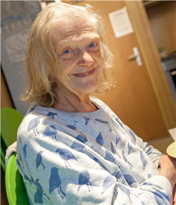 Eine Seniorin sitzt auf einem grünen Stuhl und blickt lächelnd in die Kamera