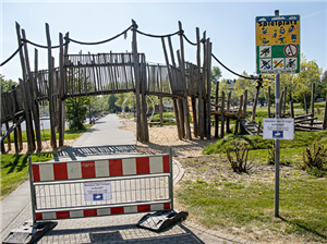 Ein Spielplatz mit einer großen Holzbrücke, dessen Eingang mit einer Absperrschranke abgesperrt ist. Auf dem Foto ist auch das Hinweisschild des Spielplatzes zu sehen.