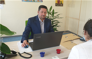 Samer Qarqash sitzt an seinem Arbeitsplatz vor einem Laptop und guckt in die Kamera. Ihm gegenüber sitzt eine Klientin mit Schutzmaske.