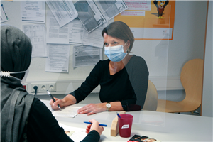 Eine Beraterin der Caritas in Geldern sitzt an Ihrem Arbeitsplatz und spricht mit einer Klientin. Beide tragen eine Maske und werden durch eine Plexiglasscheibe getrennt.