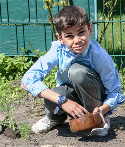 Eine Junge kniet mit einer Holzschüssel in einem Garten, sät Pflanzensamen aus und blickt dabei in die Kamera