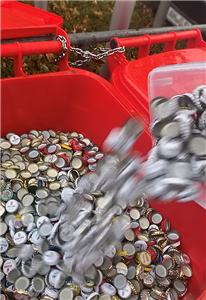 Eine Person schüttet Kronkorken von einer Kunststoffbox in eine rote Abfalltonne, die mit einer großen Zahl an Kronkorken gefüllt ist