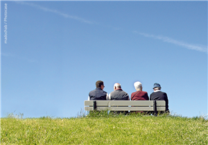Vier Senior/innen sitzen an einer Wiese vor einem wolkenlosen Himmel auf einer Bank