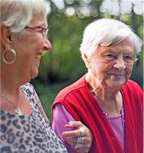 Eine weibliche Betreuungskraft geht mit einer Seniorin spazieren. Dabei hat sie einen Arm bei der Seniorin untergehakt und lächelt sie an.