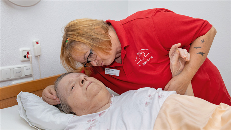 Eine Pflegerin des St. Josefshauses in Witten beugt sich zu einer in einem Pflegebett liegenden Seniorin runter und hält dabei ihren linken Arm