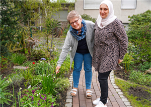 Annegret Stöcker und Ramia Al Shoueb stehen in einem Garten auf einem Steinweg zusammen und lächeln in die Kamera