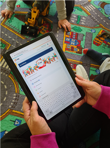 Eine Frau sitzt auf einem Spielteppich und hält ein Tablet, auf dem die Isy-App zu sehen ist, in der Hand. Neben ihr spielt ein Kind.
