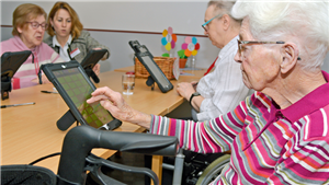 Drei Seniorinnen sitzen ein einer Tischreihe vor Tablets. Eine weitere Frau sitzt daneben und unterstützt bei der Anwendung.