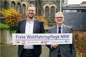 Christian Woltering und Hartmut Krabs-Höhler stehen vor einem Kirchengebäude und halten lächelnd ein Banner der LAG Freie Wohlfahrtspflege NRW nach oben