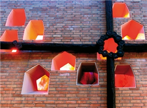 Eine Altar-Fensterwand mit einem großen schwarzen Kreuz und zehn kleinen Fenstern durch die Sonnenlicht reinfällt. Die Wand besteht aus roten Ziegelsteinen.