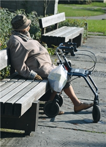 Alte Frau sitzt neben ihrem Rollator auf einer Parkbank