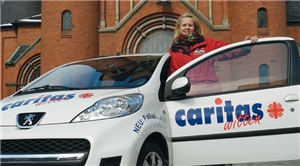Katrin Piorunek steht hinter der geöffneten Tür ihres weißen Caritas-Autos, dass vor einer Kirche steht