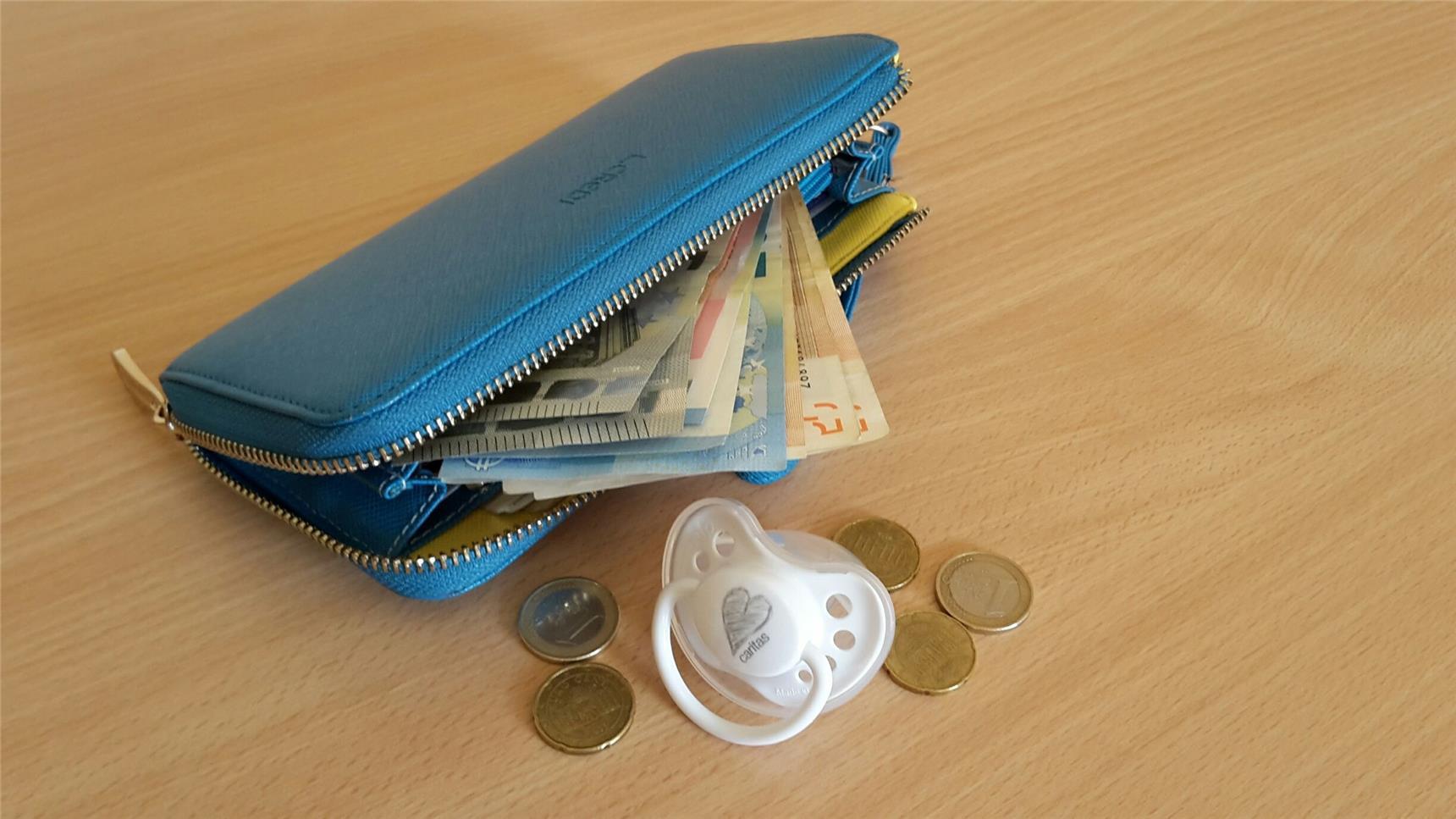 Blaue Geldbörse mit mehreren Scheinen und Münzen und ein Babyschnuller mit einem aufgedrucktem Herz und dem Schriftzug "caritas"