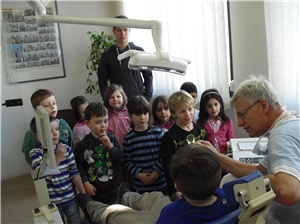 Ein Kind sitzt aufdem Behandlungsstuhl, der Zahnarzt ist neben ihm und erklärt ewas. Eine Gruppe mit Jungen und Mädchen steht um die beiden herum. Alle hören aufmerksam zu.