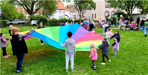 Erwachsene und Kinder spielen in einem Park mit einem regenbogenfarbenen Schwungtuch, im Hintergrund Menschen an Tischen bei gemeinsamen Frühstück