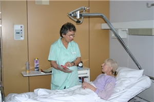 Frau mit grünem Kittel steht am Krankenbett einer älteren Frau.