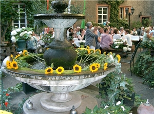 Ehrenamtstag Im Vordergrund ein Brunnen mit Sonnenblumen geschmückt. Im Hintergrund mehrere Personen an Tischen.