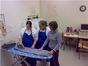 Zwei Mädchen und eine Betreuerin stehen hinter einem Bügelbrett und bügeln.