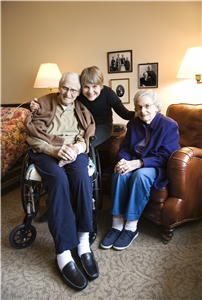 Eine Frau umarmt einen älteren Mann im Rollstuhl und eine ältere Frau, die neben ihm sitzt