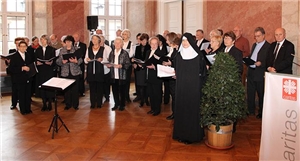 In der Ecke des Raumes im Schloss hat sich der Projektchor aufgestellt. Alle tragen festliche Kleidung. Die Leiterin, eine Ordensschwester steht rechts vor den Sängern.