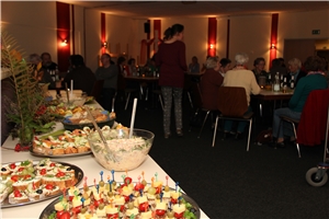 Auf diesem Bild sind mehrere Menschen abgebildet. Diese sitzen am Tisch. Im Vordergrund steht ein Buffet mit Essen.