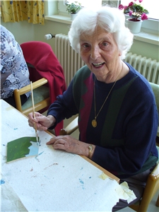 Eine ältere Damen, mit Pinsel in der Hand und einem Blatt Papier vor sich, malt. Sie schaut beim Fotografieren hoch und blickt direkt in die Kamera. 