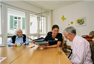 Praktikantin mit Senioren Eine junge Frau sitzt mit einer älteren Frau (links von ihr) und einem älteren Mann (rechts von ihr) am Tisch und hat einen Bildband in der Hand. Sie hat gerade in dem Buch eine Stelle gefunden, worüber sie und der ältere Mann lachen.
