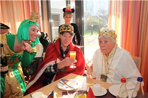 Prinzenpaar mit Sektgläsern sitzt am Tisch, daneben junge Frau in grünem Kleid mit Krone