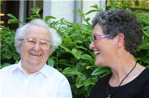 Eine Mitarbeiterin und ein Bewohner stehen vor dem Gebäude vor einer Hecke, beide lachen. Eine Mitarbeiterin und ein Bewohner stehen vor dem Gebäude vor einer Hecke, beide lachen.