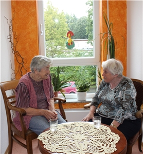 Zwei Bewohnerinnen sitzen an einem Tisch mit einer beigefarbenen Häkeldecke in einer gemütlichen Sitzecke und plaudern. 1.4. Wohnen und Leben Sitzecke im Haus