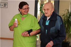 Eine Mitarbeiterin stützt einen Bewohner beim Gehen. Sie lächelt ihm zu. Eine Mitarbeiterin stützt einen Bewohner beim Gehen. Sie lächelt ihm zu.