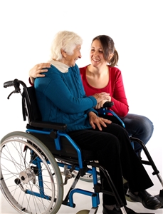 Eine Mitarbeiterin der Altenpflege legt den Arm um eine ältere Frau im Rollstuhl und hält ihre Hand. 